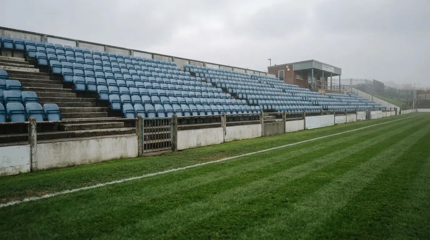 Rzędy pustych plastikowych krzesełek małego stadionu widziane z poziomu murawy pod pochmurnym niebem.