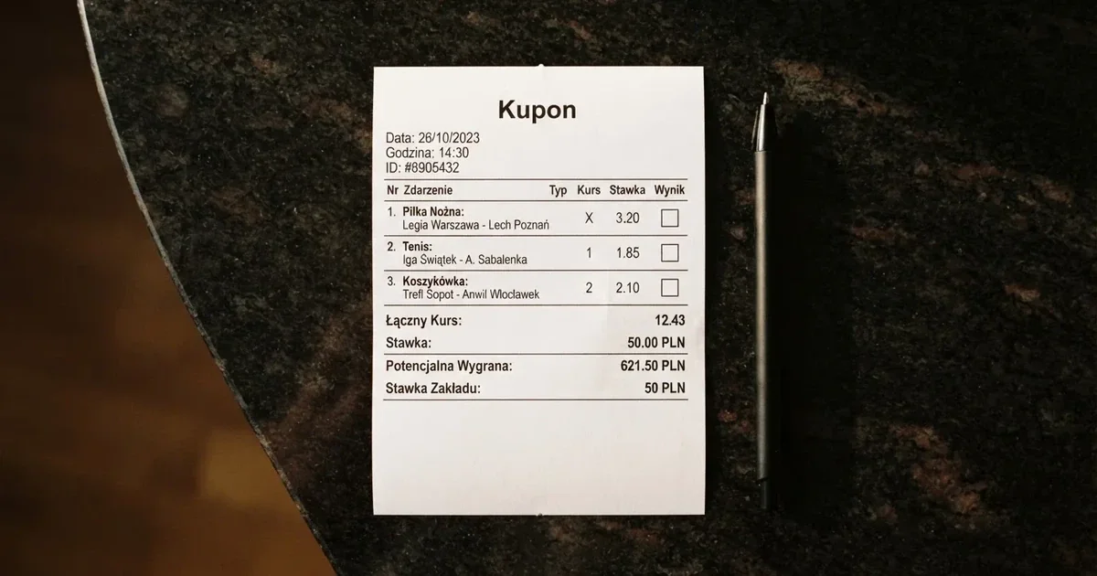 Biały papierowy kupon z pustymi polami leży na ciemnym blacie obok długopisu, widok z góry.