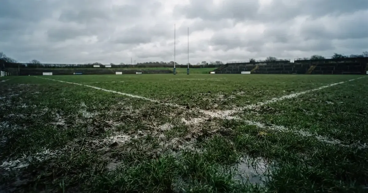 Boisko do rugby ze słupami bramki w oddali pod pochmurnym niebem, mokra trawa i ślady butów.