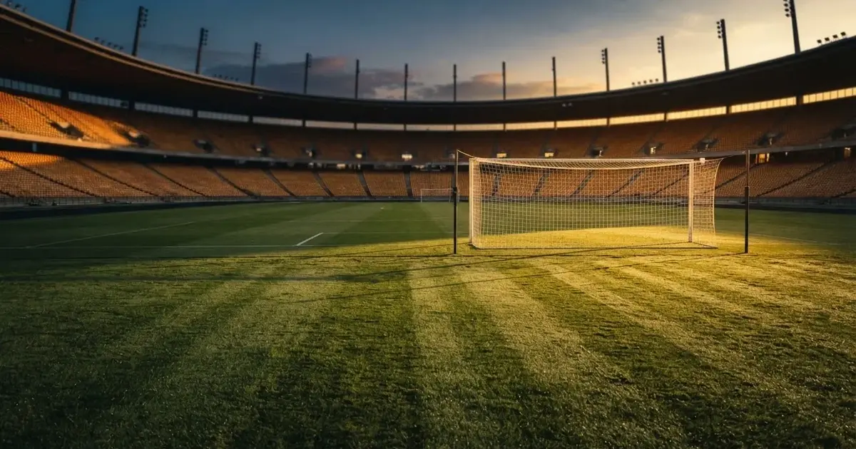 Murawa stadionu o zachodzie słońca z długimi cieniami słupków bramki, trybuny w ciepłym świetle.