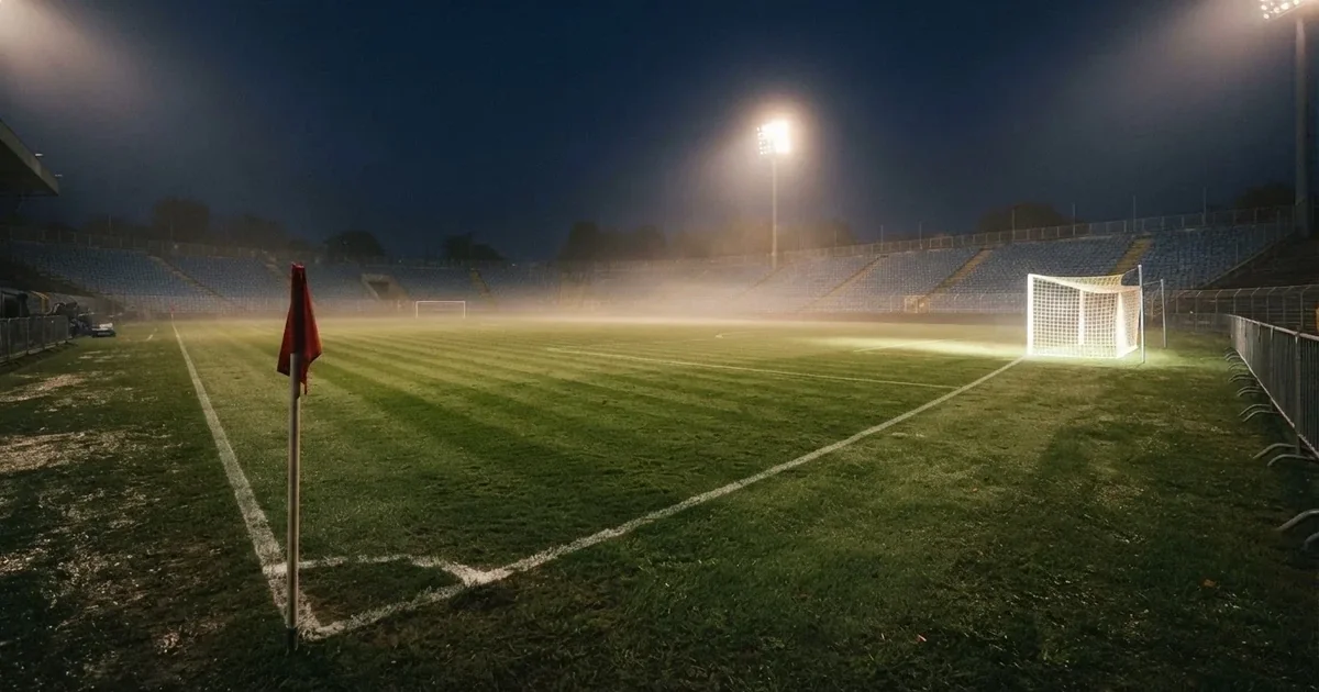 Pusta murawa stadionu miejskiego w chłodny jesienny wieczór, siatka bramki świeci w świetle reflektorów, nad boiskiem wisi mgła.